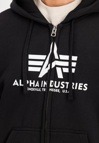 Crna zip-up dukserica od pamuka s prednjim grafičkim prikazom "Alpha Industries" u bijelom tekstu i stiliziranim logotipom. Sadrži vezice i džepove.