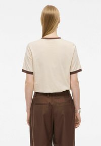 T-shirt beige a maniche corte con rifiniture marroni a coste, che presenta una silhouette rilassata. Abbinato a pantaloni marroni a vita alta.