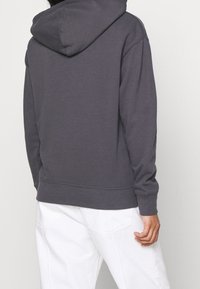 Personne portant un sweat à capuche gris foncé et un pantalon blanc, vue de dos avec une posture détendue et une main visible.