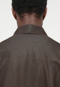 Vue arrière d'une personne aux cheveux courts et bouclés portant une veste en cuir marron foncé avec le nom de la marque embossé sur la partie supérieure du dos.