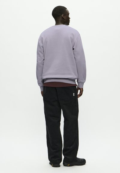 Sweat-shirt lilas avec des poignets côtelés, associé à un pantalon en velours côtelé noir avec des poches. Le modèle se tient de dos contre un fond uni.