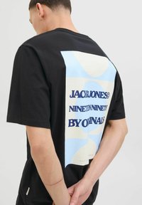 Sort bomulds T-shirt med et stort grafisk print i blå og crème på bagsiden, der viser tekst og abstrakte former. Kortærmet.