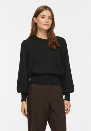 Jeune femme aux longs cheveux bruns portant un pull noir à manches longues et un pantalon marron, debout devant un fond uni.