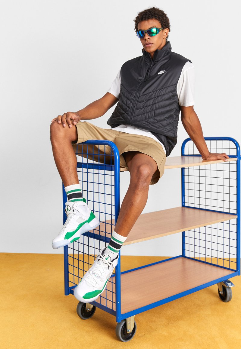 Schwarze gesteppte Weste über einem weißen Hemd, beigefarbene Shorts, weiße und grüne Turnschuhe, sitzend auf einem blauen Rollregal mit Holzregalen.