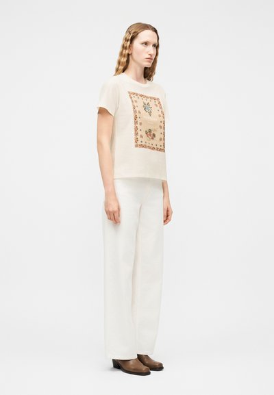 Γυναίκα με κυματιστά ξανθά μαλλιά στέκεται φορώντας ένα μπεζ μπλουζάκι με λουλουδάτο σχέδιο, άσπρο παντελόνι με φαρδιά μπατζάκια και καφέ μπότες με τακούνι, μπροστά σε λευκό φόντο.