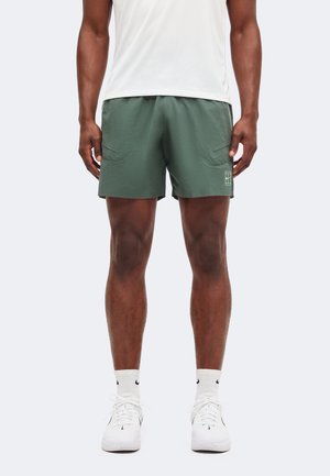 Mand iført mørkegrønne atletikshorts med forlommer, hvide sportssokker, hvide sneakers og en hvid skjorte, stående mod en ensfarvet baggrund.