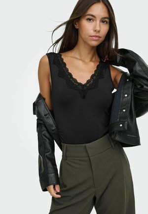 Femme portant un haut sans manches noir avec bordure en dentelle, un pantalon taille haute vert foncé et une veste en cuir noire posée sur ses épaules.