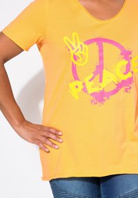 T-shirt jaune à manches courtes avec un symbole de paix rose et un graphique "PEACE" en néon jaune avec des détails éclaboussés. Tissu doux et léger.