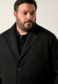 Cappotto di lana nera con colletto a rever, chiusura frontale a singolo bottone e tasche laterali, indossato sopra una polo nera con colletto bianco.