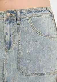 Jasnoniebieska spódnica dżinsowa z efektem przetarcia, widocznym przeszyciem, zamkiem błyskawicznym z przodu oraz kieszeniami. Tkanina ma wyblakły kolor.