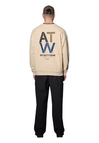 Kremowa bluza z dużym niebieskim i brązowym napisem na plecach, zestawiona z czarnymi spodniami cargo. Napis zawiera "OFF DUTY GEAR" oraz "HALO."