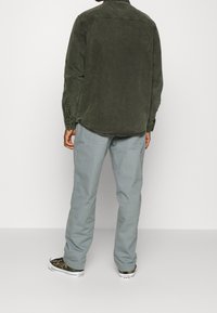 Zielona bawełniana koszula o luźnym fasonie, z długimi rękawami i zaokrąglonym dołem, połączona z jasnoszarymi spodniami i sneakersami w kamuflażu.