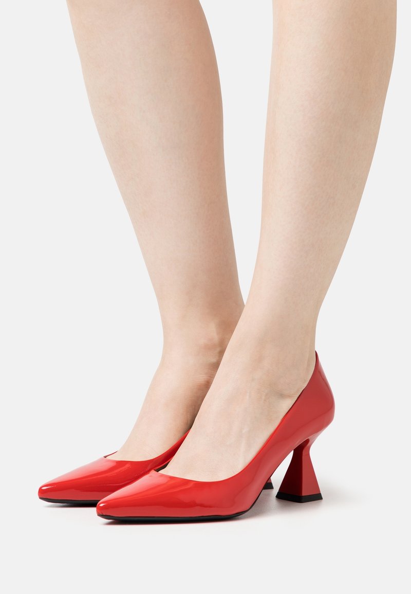 Jambes nues portant des chaussures à talons hauts rouges brillantes avec un talon bloc évasé sur un fond clair uni.