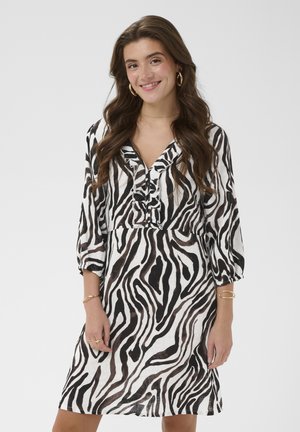 Kobieta o długich brązowych włosach ubrana w sukienkę w czarno-biały wzór zebry z falbanami u dekoltu w kształcie litery V i rękawami do połowy przedramienia, uśmiecha się na tle jednolitego tła.