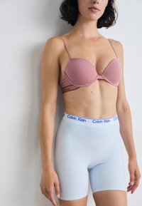 Persona che indossa un reggiseno rosa e biancheria intima azzurra chiara, con uno sfondo di muro bianco, che mostra uno stile moderno e casual.