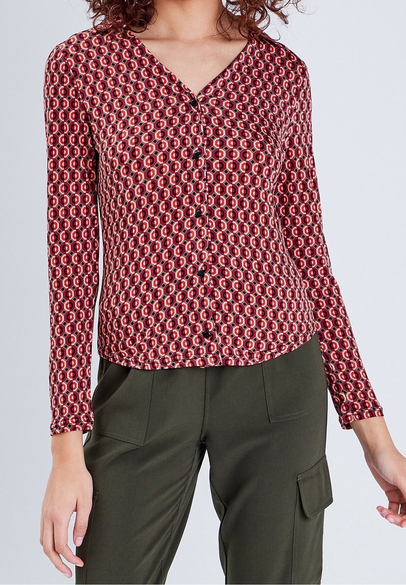 Femme portant un chemisier à manches longues, à motif géométrique rouge et rose, boutonné, et un pantalon cargo vert olive foncé.