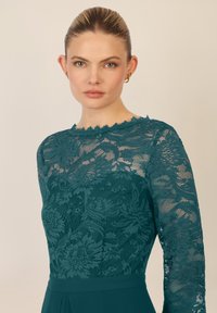 Top in pizzo turchese con maniche lunghe, caratterizzato da motivi floreali e scollatura ondulata. Tessuto liscio sotto con una silhouette elegante.