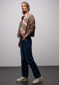 Veste bomber marron avec poignets côtelés, jean bleu marine à jambes larges, et baskets de couleur claire. Enveloppée d'une écharpe pastel à motifs.