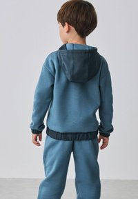 Blauwe trainingsoutfit bestaande uit een hoodie met een donkere rand en elastische manchetten, gecombineerd met bijpassende broeken. Zachte stof met een gladde textuur.
