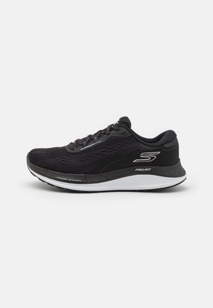Zapatilla deportiva negra con parte superior de malla, punta redondeada, entresuela blanca en contraste y suela de goma negra. Presenta cordones y detalle de logo en el lateral.