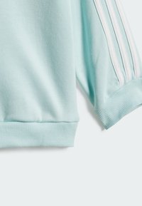 Sudadera azul claro con puños y dobladillo acanalados, que presenta tres rayas blancas a lo largo de la manga. Textura suave con un diseño deportivo.