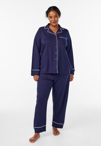 Set di pigiama blu navy con maniche lunghe e apertura a bottoni. Presenta rifiniture bianche lungo i bordi e una tasca sul petto. Tessuto morbido e confortevole.