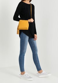 Musta pitkähihainen paita, siniset skinny-farkut, valkoiset tennarit ja keltainen neliönmuotoinen olkalaukku, jossa on hapsu koristeena. Puhdas, moderni estetiikka.