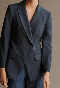 Tmavě modrý přizpůsobený sako s klopovým límečkem, zapínání na jeden knoflík, dvě přední kapsy a ostré, strukturované linie. Vyrobeno z texturované látky.