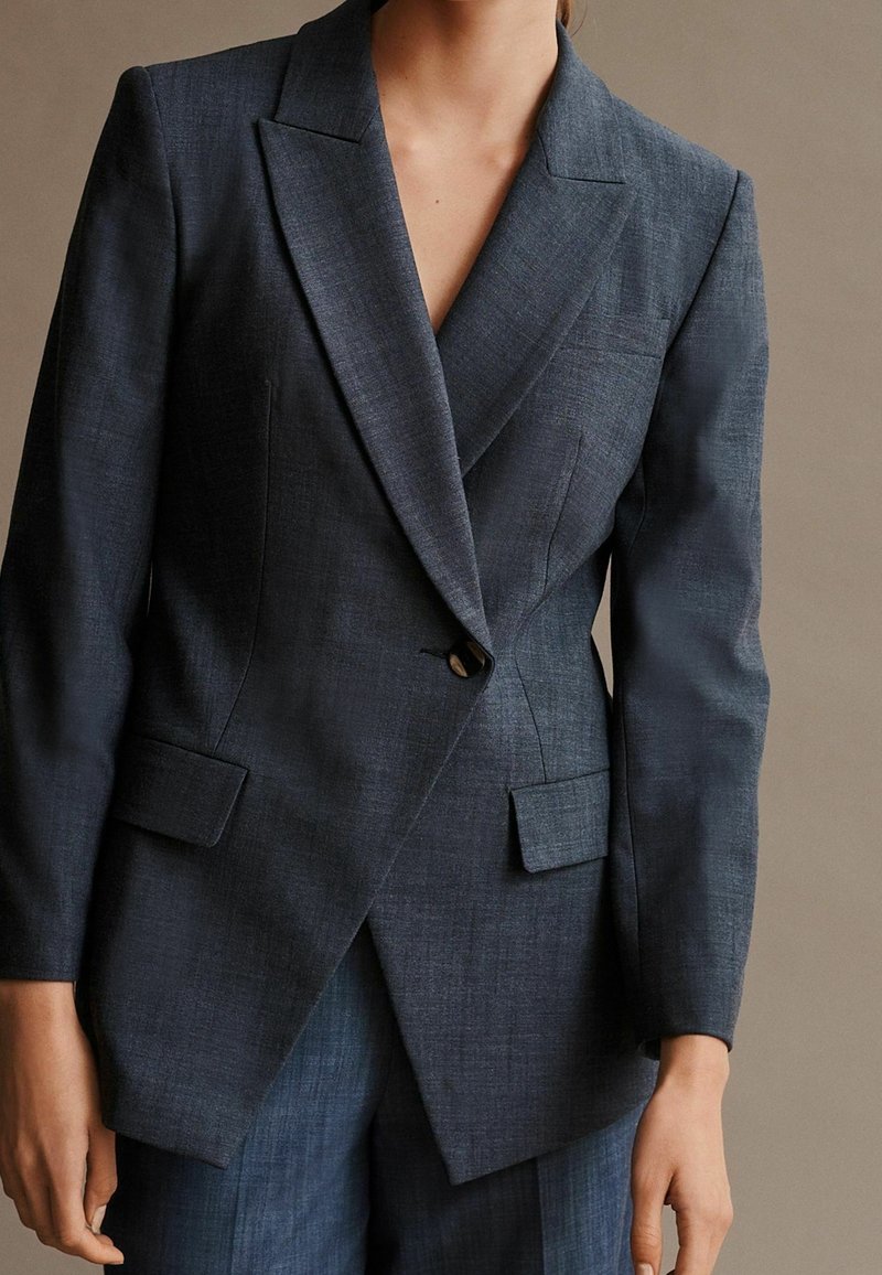 Tmavě modrý přizpůsobený sako s klopovým límečkem, zapínání na jeden knoflík, dvě přední kapsy a ostré, strukturované linie. Vyrobeno z texturované látky.