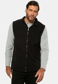 Schwarze Fleece-Weste mit einem hohen Kragen, durchgehendem Reißverschluss und zwei seitlichen Taschen. Getragen über einem hellgrauen Langarmhemd.