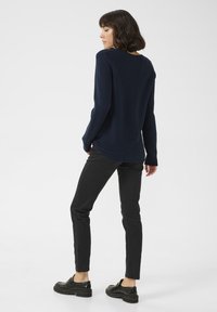 Marineblaues, geripptes Langarmoberteil, schwarze, eng anliegende Hose und schwarze klobige Schuhe. Modell steht mit dem Rücken zur Kamera und präsentiert das Rückendesign sowie die Outfitkombination.