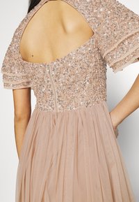 Vestido de noche con cuentas y mangas cortas en forma de volantes, espalda descubierta y falda de tul en rosa suave. Presenta un cierre central con cremallera en la parte posterior.