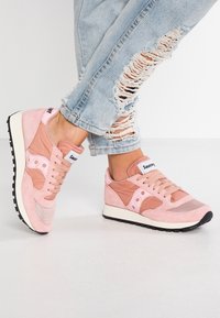 Baskets roses portées avec un jean bleu clair déchiré, les pieds croisés sur un fond blanc uni.