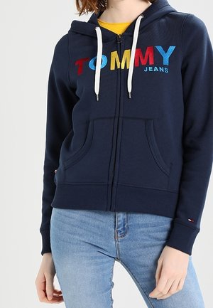 Persoon draagt een marineblauwe hoodie met ritssluiting en kleurrijke "TOMMY JEANS" tekst, gecombineerd met lichtblauwe spijkerbroek tegen een effen achtergrond.
