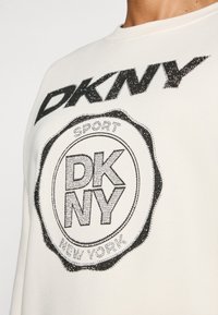 Felpa color crema chiara con logo DKNY in strass neri e design a badge circolare, con la scritta "SPORT" e "NEW YORK" stampata sotto.