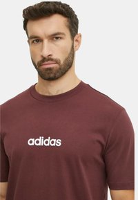 T-shirt di cotone color marsala con collo rotondo e maniche corte. Nota il logo bianco "adidas" sul petto. Texture liscia.