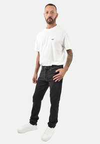 T-shirt bianca a maniche corte con logo, abbinata a jeans neri. Il modello indossa sneaker bianche. Lo sfondo neutro mette in risalto i dettagli dell'outfit.