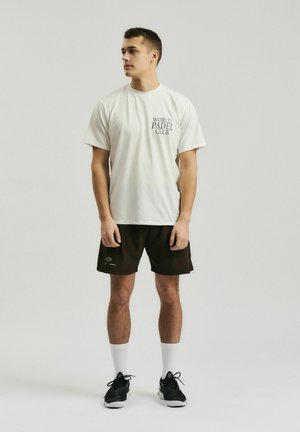 Ung mand stående iført en hvid World Padel Club t-shirt, sorte shorts, hvide sokker og sorte sportssko på en ensfarvet baggrund.
