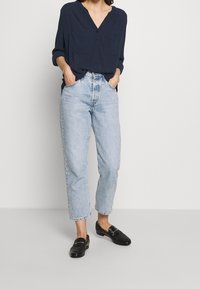 Donna che indossa una blusa blu navy con bottoni, jeans a vita alta blu chiara e mocassini in pelle nera, in piedi con le mani nelle tasche.