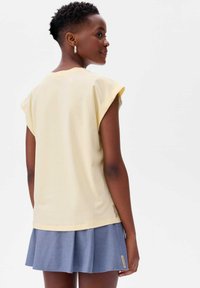 Lichtgele top met korte mouwen en omgeslagen manchetten, gecombineerd met een geplooide denim minirok. Huidskleur is middelbruin.