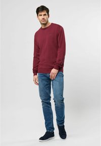 Ein Mann steht vor einem schlichten, hellgrauen Hintergrund und trägt einen bordeauxfarbenen Pullover, blaue Jeans und schwarze Sneakers.
