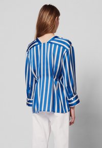 Modrá a bílá pruhovaná blůza z hladké tkaniny s V-zadním výstřihem, dlouhými rukávy a zapínacími manžetami. Přiléhavý střih s bočními rozparky.