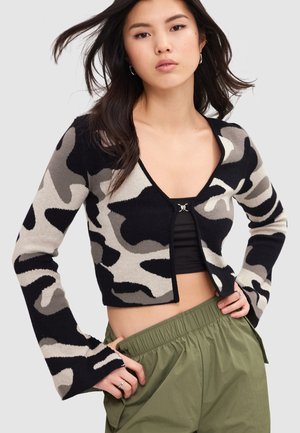 Mujer joven con un cárdigan corto camuflado en negro, gris y beige, top corto negro y pantalones verde oliva con las manos en las caderas.