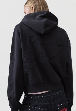 Blonde person wearing black hoodie with stitched star patches on the back and sleeves, paired with black studded jeans with red lip design.