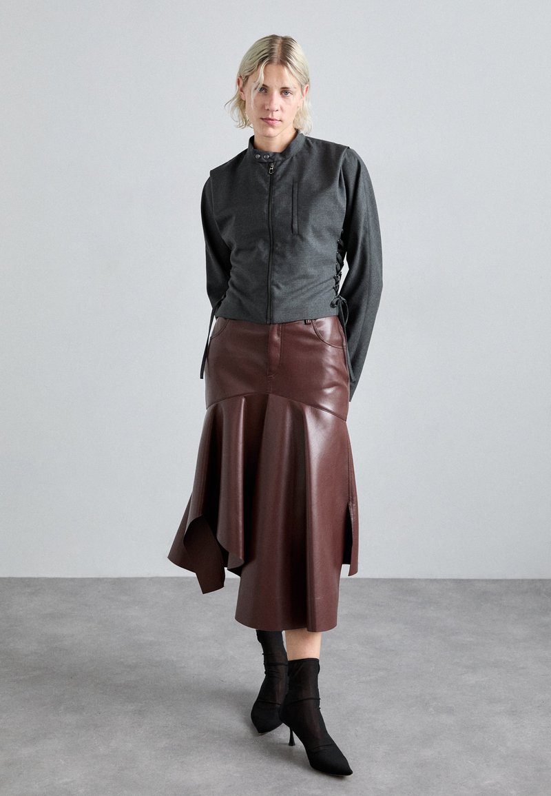 Giacca grigia corta con zip frontale, maniche lunghe e legami laterali. Gonna asimmetrica in finta pelle marrone con orlo svasato. Stivali neri con tacco alto.