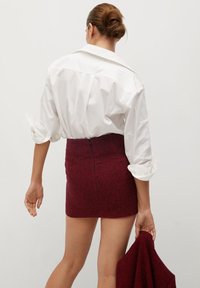 Camicia bianca oversize con maniche arrotolate, abbinata a una gonna corta borgogna strutturata. La gonna presenta una chiusura con zip sul retro.