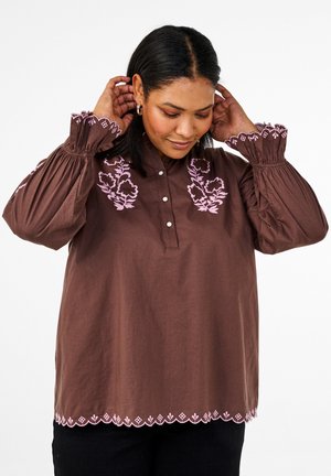 Blusa in cotone marrone con maniche lunghe a sbuffo, caratterizzata da ricami floreali rosa sul petto e orlo ondulato. Chiusura frontale con bottoni.