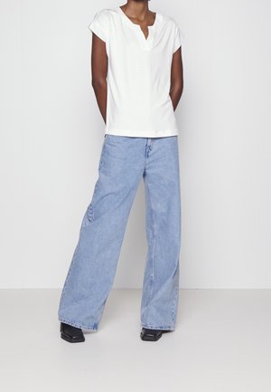 Person iført en hvid kortærmet top med en notched halsudskæring og lyseblå viddejeans med sorte sko, stående mod en ensfarvet væg.