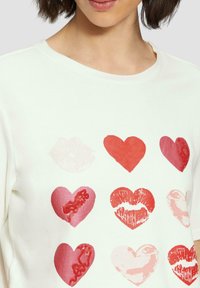Witte katoenen t-shirt met een raster van rode en roze hartafdrukken, sommige met lipafdrukken, variërend in tinten en texturen.