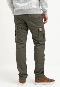 Olivefarbene Cargo-Hosen mit mehreren seitlichen Taschen und einem Carhartt-Logo-Patch. Kombiniert mit braunen Sneakers vor einem hellen Hintergrund.